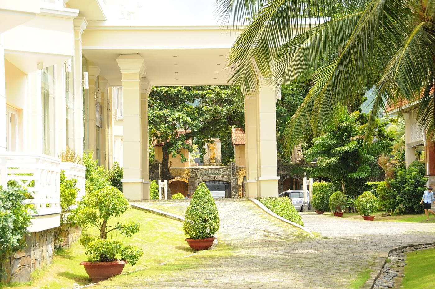 Подъезд к главному корпусу Saigon Côn Đảo Resort: колонны, мощёная дорожка, пальмы и ворота тюрьмы Phú Sơn напротив