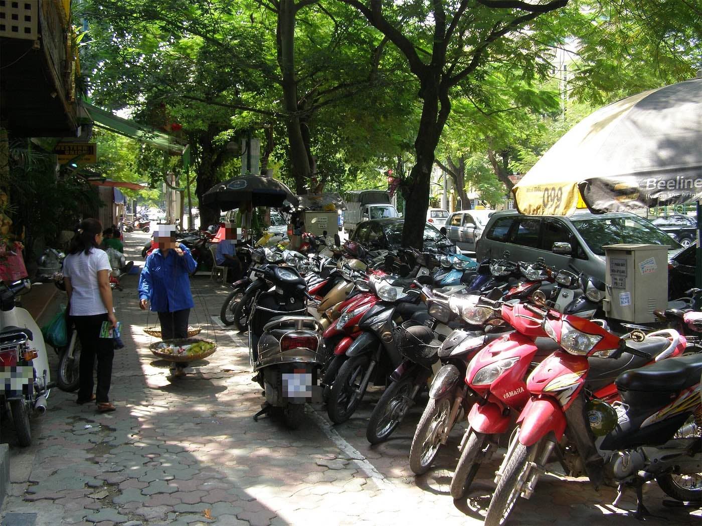 Motorbikes parked on a tree-lined sidewalk in Hanoi next to a fruit vendor