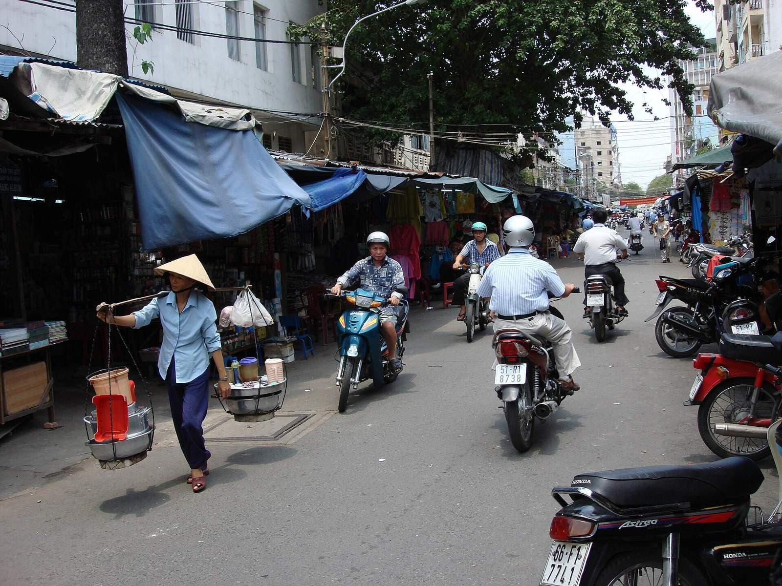 A Saigon market street with motorbikes, awnings and a vendor carrying a shoulder pole