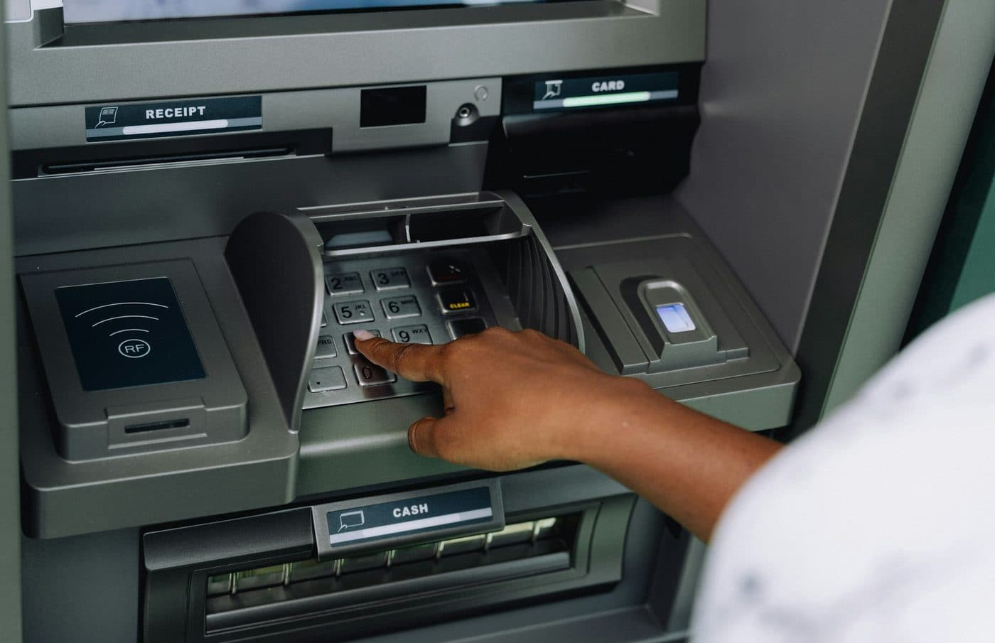 Рука вводит PIN на клавиатуре банкомата под защитным козырьком