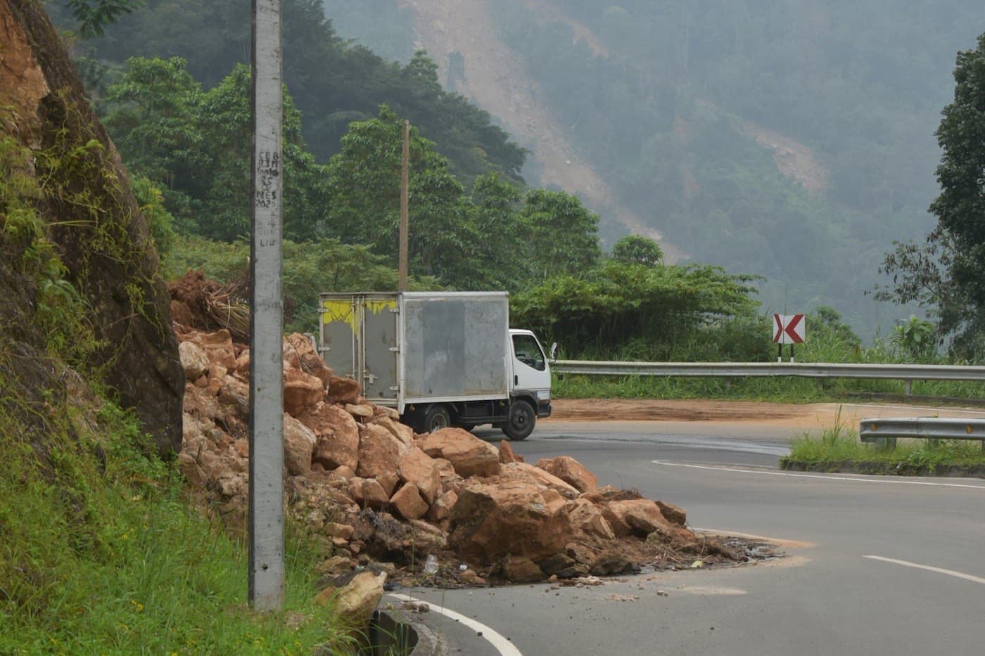 Landslide rocks on a bend of a mountain road with a truck behind and scarred slopes